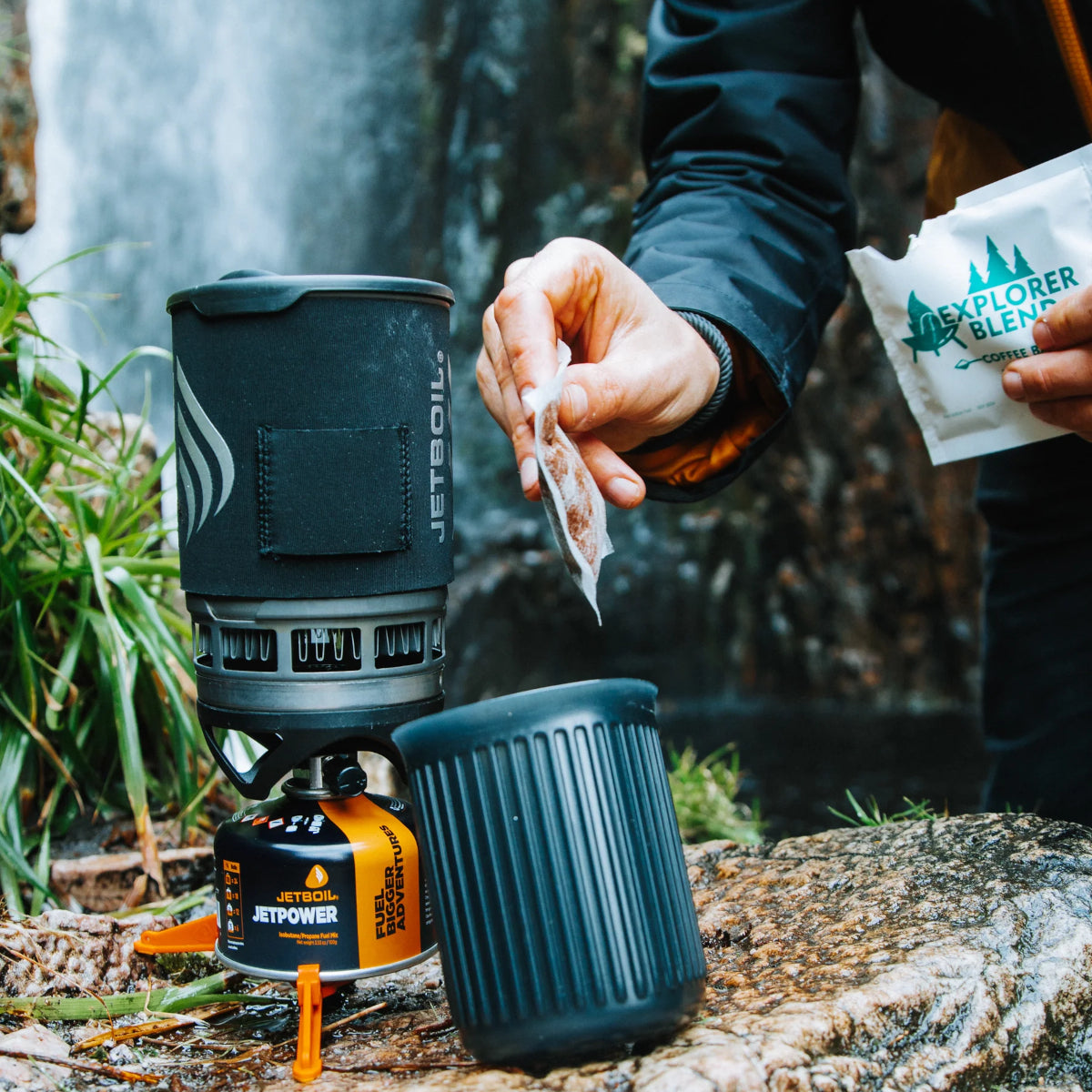 Person preparing a hot beverage using a Jetboil stove outdoors with a waterfall in the background.