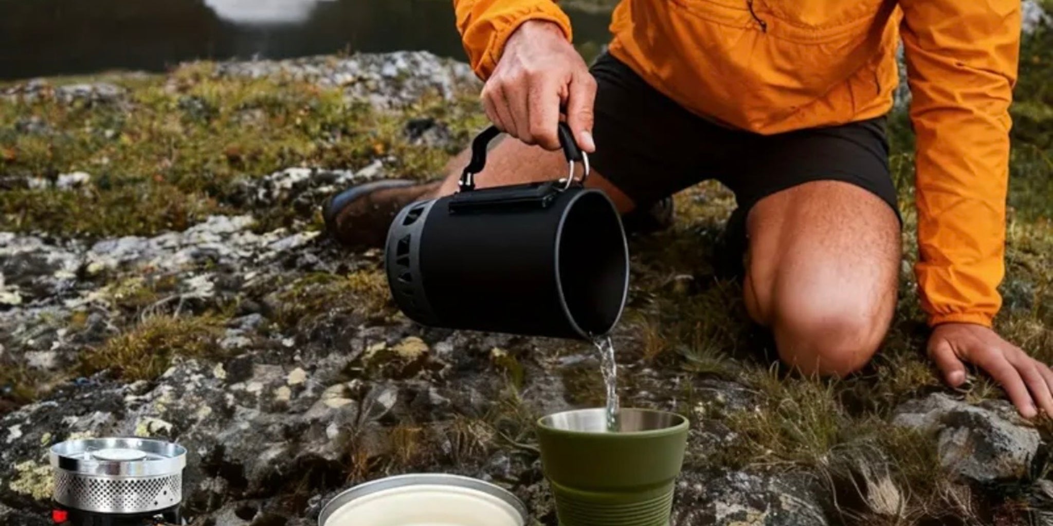Person in orange jacket pouring water from a black pot into a green cup outdoors.
