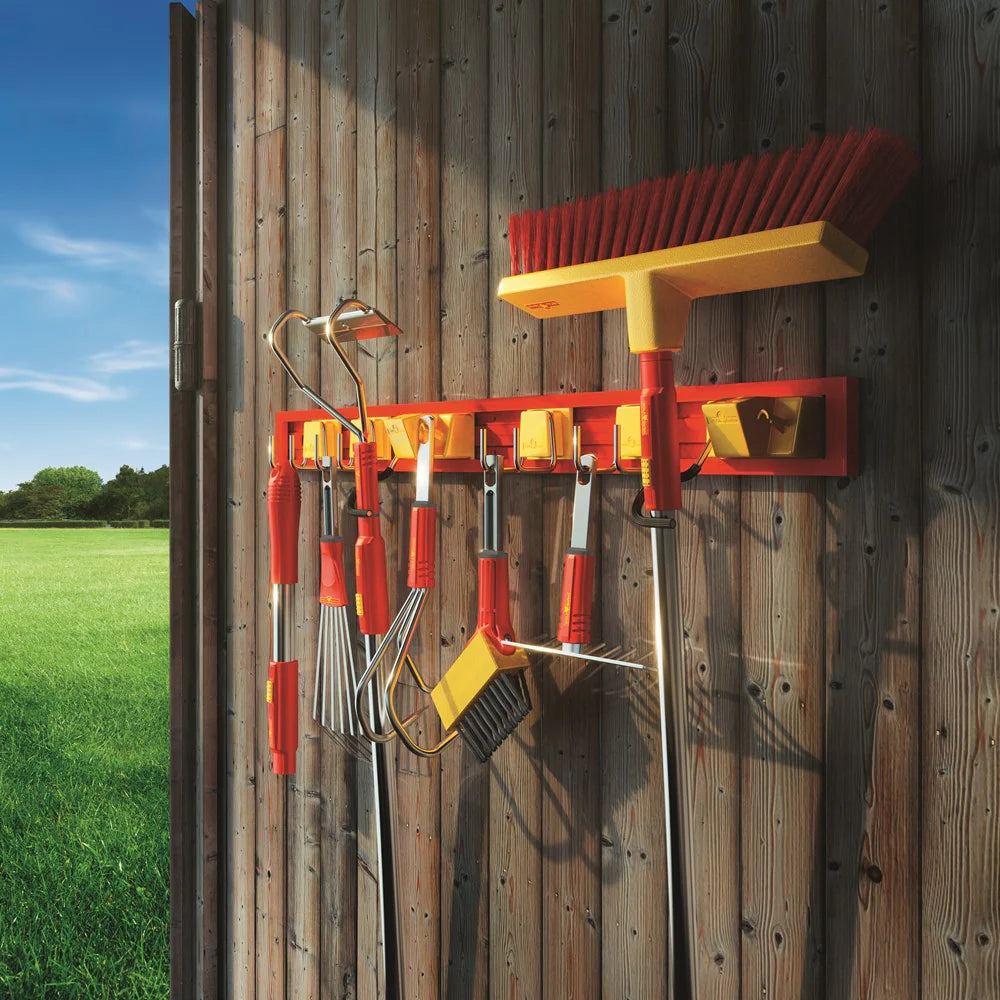 Garden tools including a broom and rakes on a wall-mounted holder against a wooden fence with a grassy field and blue sky in the background.